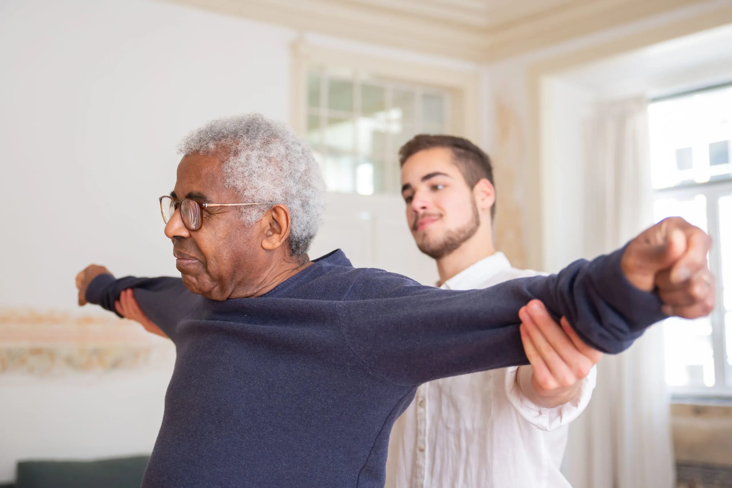 An older man stretches his arms with a helper at home