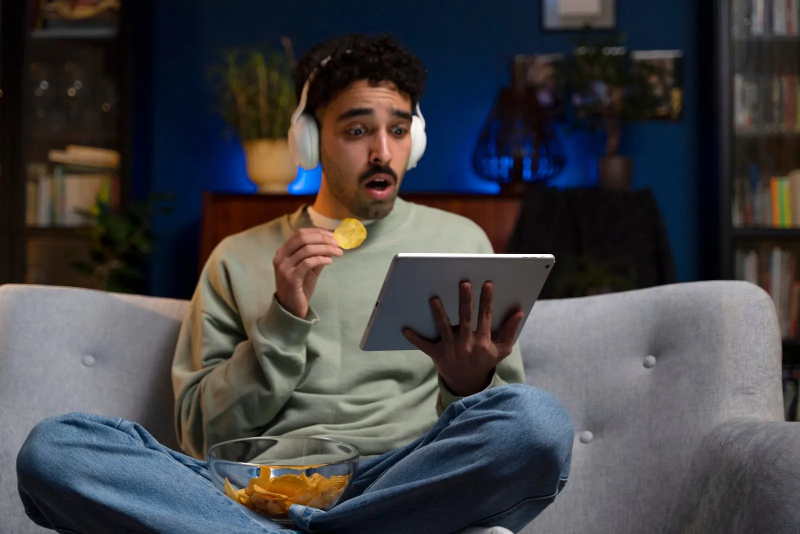A young man sitting on a couch, wearing headphones and holding a tablet while eating chips. He appears surprised or entertained, highlighting modern screen time and digital media consumption habits.